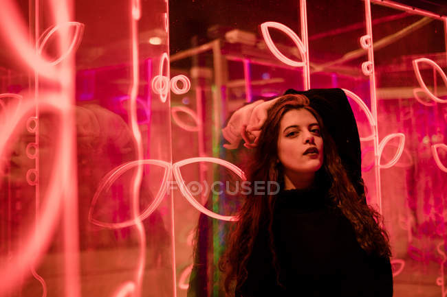 Pretty pensive young woman in casual wear looking in camera among neon signs at city street — Stock Photo