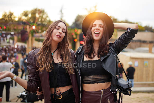 Long haired fashion friends in leather jacket and hat dancing raising hands with two fingers up and looking at camera on festival in sunny day — Stock Photo