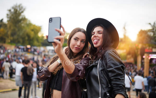 Очаровательные веселые друзья в черной шляпе веселятся гримасом и делают селфи на мобильном телефоне в яркий день на фестивале — стоковое фото