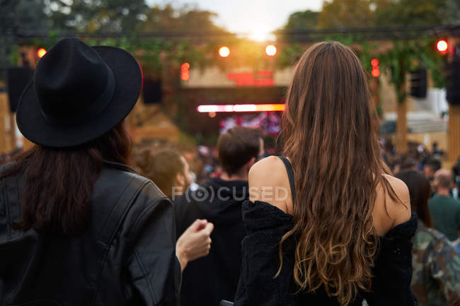 Back view of stylish girl friends in black hat taking selfie and watching photo on mobile phone in bright day at festival — Stock Photo