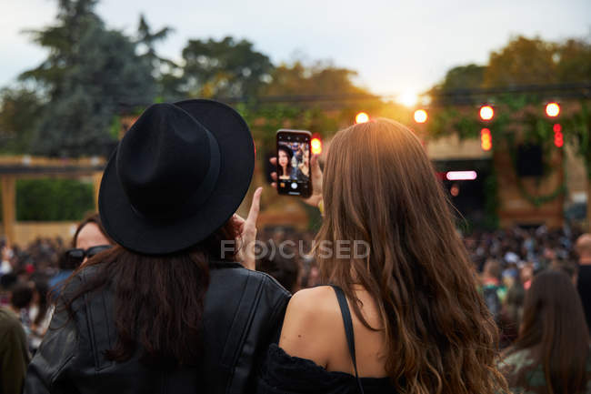 Back view of stylish girl friends in black hat taking selfie and watching photo on mobile phone in bright day at festival — Stock Photo