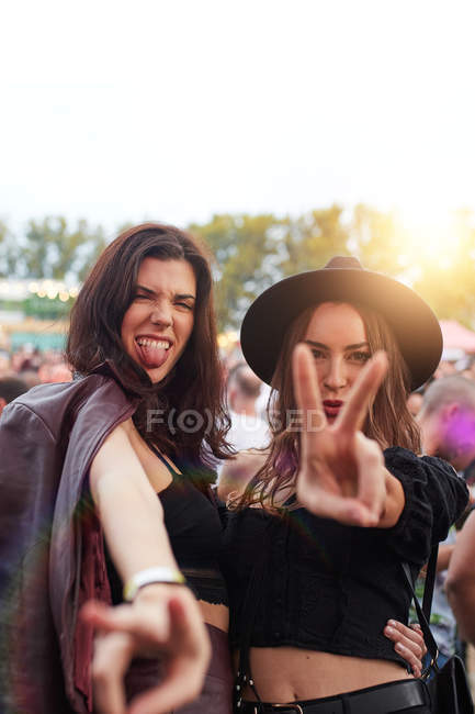 Elegantes mujeres de pelo largo en sombrero y chaqueta de cuero haciendo muecas sacando la lengua y mostrando dos dedos en la cámara en un día brillante en el festival - foto de stock