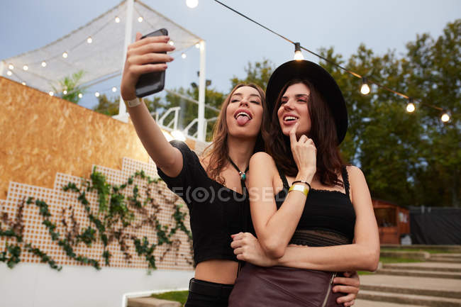 Stylische, gut gelaunte Freunde mit schwarzem Hut umarmen sich und machen bei strahlendem Sonnenschein ein Selfie auf dem Handy in der festlich geschmückten Arena — Stockfoto