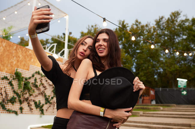 Amigos alegres elegantes no chapéu preto que abraça e toma a selfie no telefone celular no dia brilhante na arena decorada no festival — Fotografia de Stock