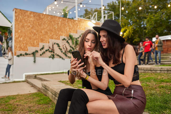 Приятные веселые друзья в черной шляпе весело смотрят фото на мобильном телефоне, сидя на зеленой лужайке рядом с украшенной сценой в яркий день на фестивале — стоковое фото