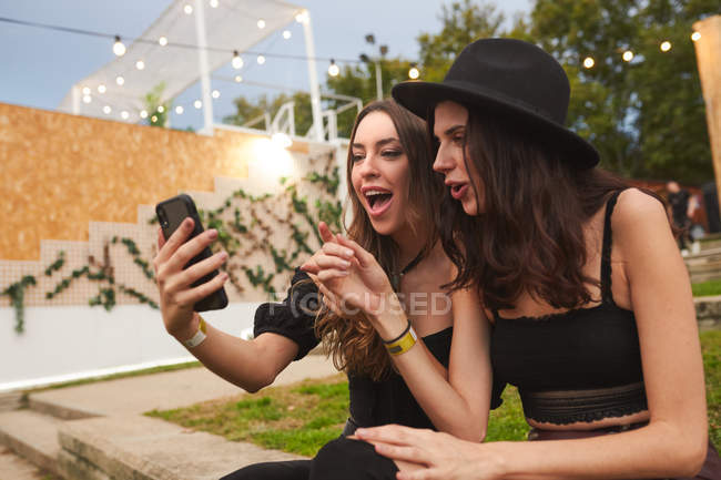 Amigos en sombrero negro que se divierten viendo la foto en el teléfono móvil sentado en el césped verde cerca del escenario decorado en día brillante en el festival - foto de stock