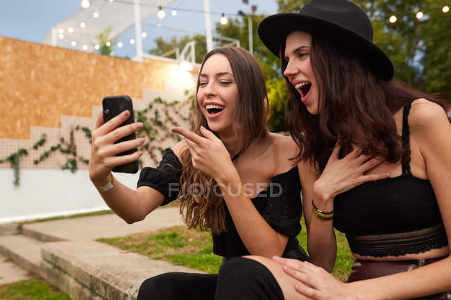 Друзья в черной шляпе весело смотрят на фото на мобильный телефон, сидя на зеленой ленточке возле украшенной сцены в яркий день на фестивале — стоковое фото