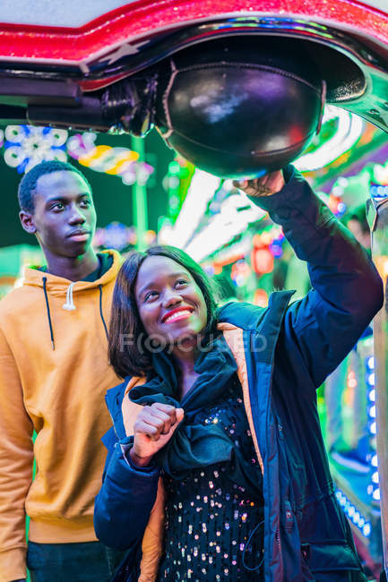 Happy black woman smiling and punching ball while playing arcade game with boyfriend at night on funfair — Stock Photo