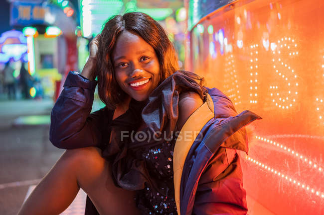 Optimistic African American female smiling and looking at camera while sitting near illuminated arcade in evening on fairground — Stock Photo