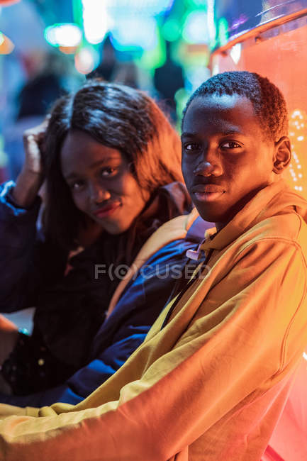 Side view of African American man looking at camera while sitting near girlfriend during date at night on funfair — Stock Photo