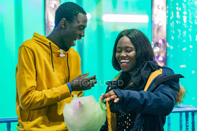 Homem e mulher afro-americanos excitados rindo e comendo algodão doce durante a data na feira — Fotografia de Stock