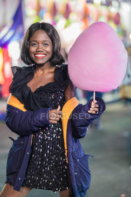 Mujer negra optimista con hilo de caramelo sonriendo y mirando a la cámara mientras se divierten en el recinto ferial por la noche - foto de stock