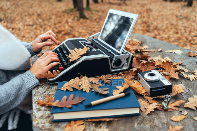 Mujer irreconocible escribiendo en máquina de escribir vintage con tableta en hojas de otoño en mesa de piedra en bosque de roble - foto de stock