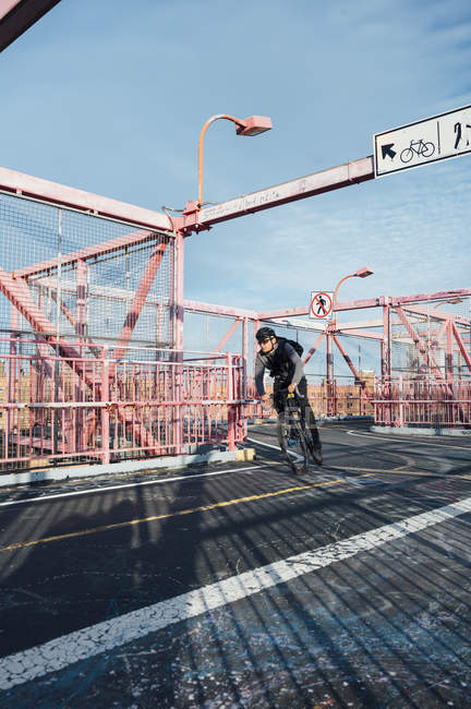 Cyclist in eyeglasses and helmet riding bike on asphalt road among red metal structure with blue sky on background in New York city — Stock Photo