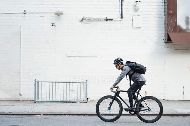 Вид збоку сучасного чоловічого велосипедиста в чорному шоломі та окулярах, що серфінгують на мобільному телефоні на фоні білої стіни — стокове фото