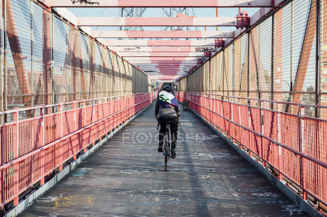 Back view of anonymous cyclist with helmet riding bike among red metal bridge structure in New York city — Stock Photo