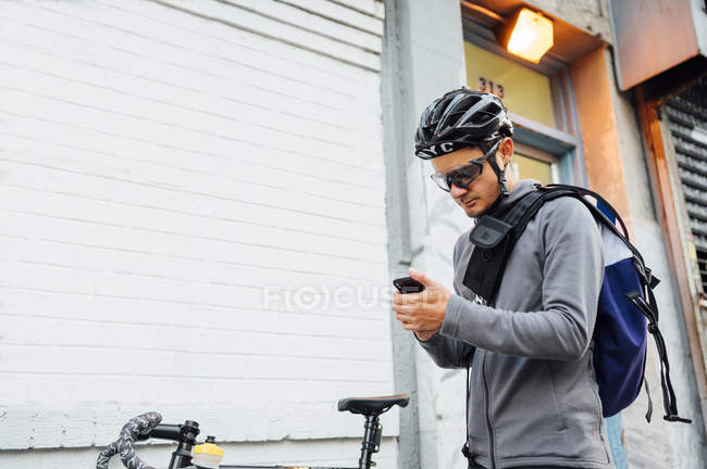 Современный мужчина велосипедист в черном шлеме и очках серфинг на мобильном телефоне на белом фоне стены — стоковое фото