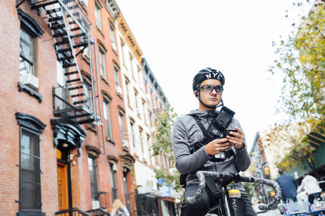 From above of male cyclist in stylish helmet and eyeglasses using mobile phone and looking away on bike at city street — Stock Photo