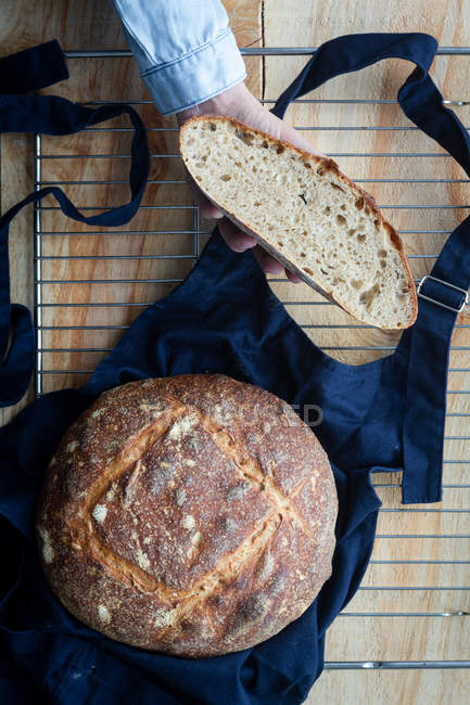 Вид сверху на руку анонимной женщины с ломтиком домашнего хлеба из теста и буханкой хлеба на фартуке на решетке печи . — стоковое фото