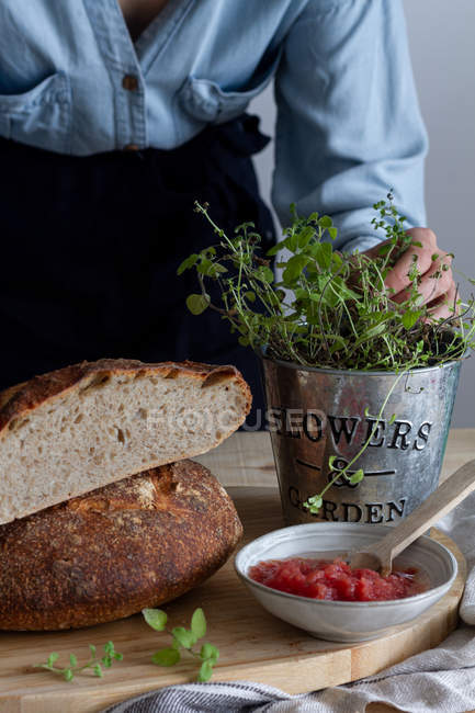 Unrecognizable female touching pot of plant near sourdough bread near red salsa sauce on wooden table — Stock Photo