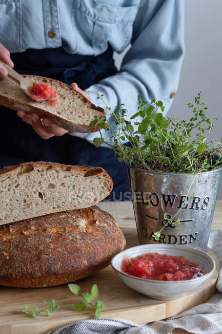 Невизначена жінка, що розкладається ложкою червоного соусу сальси на скибочці хліба на дерев'яному столі, прикрашеному зеленою рослиною — стокове фото