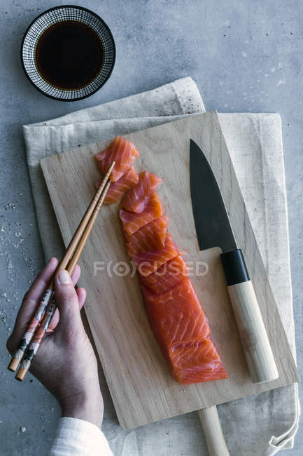 From above hand of anonymous person holding piece of salmon with chopsticks and dipping in soy sauce at served table — Stock Photo