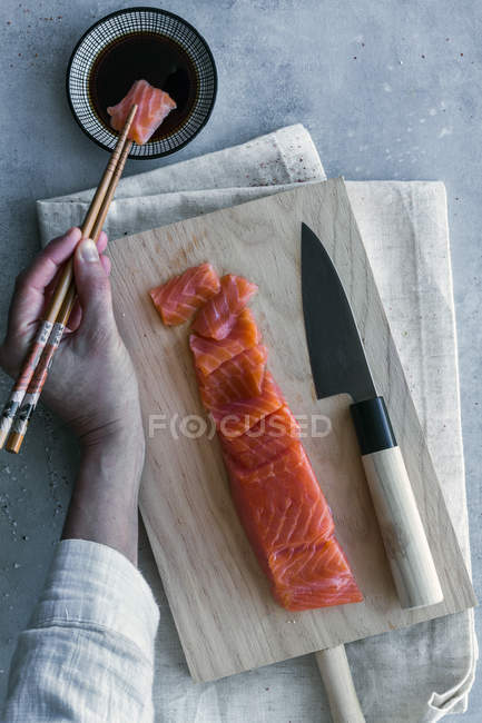 Сверху рука анонимного человека, держащего кусок лосося с палочками для еды и погружающегося в соевый соус за обслуживаемым столом — стоковое фото