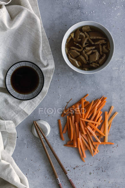 Des bâtonnets de carottes fraîches appétissants, des champignons shiitake et de la sauce soja servis sur une serviette de lin à table avec des baguettes — Photo de stock