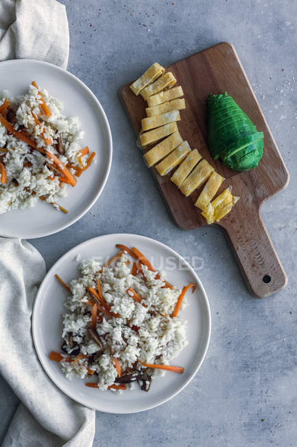 Сверху аппетитный белый рис с овощами в тарелках с яичным омлетом и разрезанным авокадо на деревянной доске — стоковое фото
