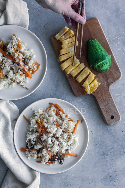 De cima mão de pessoa que toma omelete de ovo com pauzinhos da tábua de madeira com abacate de redução e chapas de arroz com verduras na mesa — Fotografia de Stock