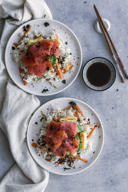 Vista superior de sabroso y apetitoso salmón en rodajas sobre arroz blanco con verduras en platos, salsa de soja y palillos en la mesa - foto de stock