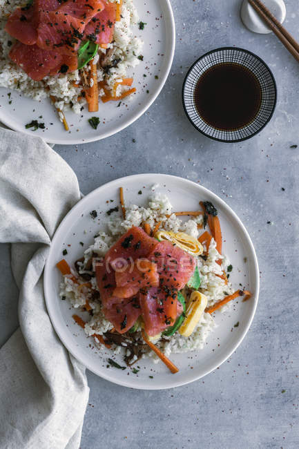 Top view of tasty appetizing sliced salmon on white rice with vegetables in plates, soy sauce and chopsticks on table — Stock Photo