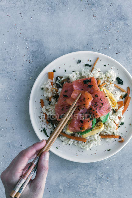 Зверху рука невизначеної людини тримає дерев'яні палички над нарізаним лососем на білому рисі з овочами в тарілці — стокове фото