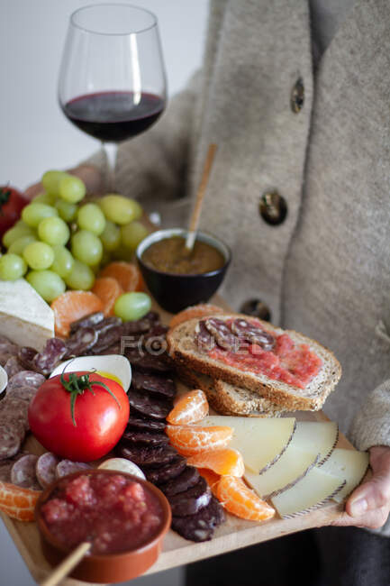 Serveur sans visage portant plateau en bois avec tranches de légumes de viande fruits et verre de vin rouge — Photo de stock