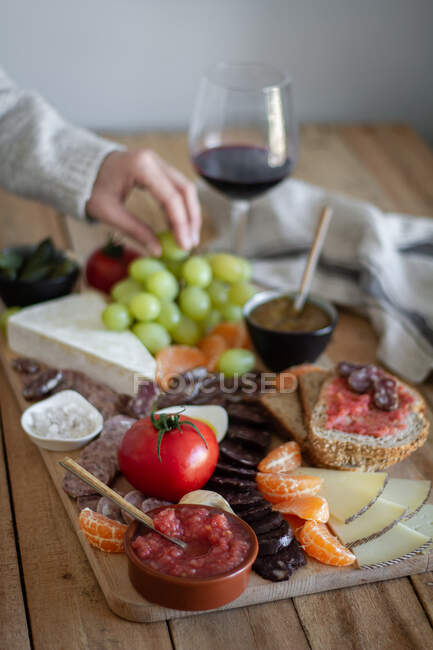From above faceless cropped person hands eating snacks from wooden tray with sliced of meat vegetables fruits and glass of red wine — Stock Photo