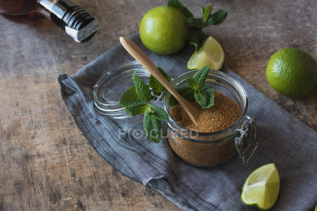 Overhead braunen Zucker in einem Glas in der Nähe von frischen Limetten und Pfefferminzblättern auf Serviette auf einem Holztisch platziert — Stockfoto