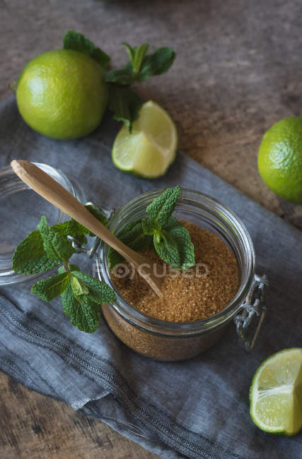 Azúcar moreno en un frasco cerca de limas frescas y hojas de menta colocadas en una servilleta sobre una mesa de madera - foto de stock