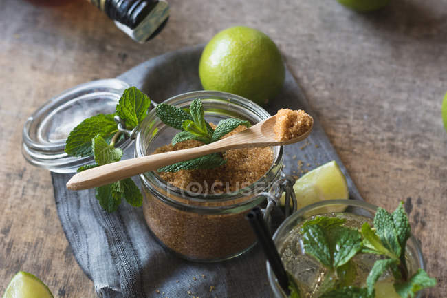 Azúcar moreno en un frasco cerca de limas frescas y hojas de menta colocadas en una servilleta sobre una mesa de madera - foto de stock