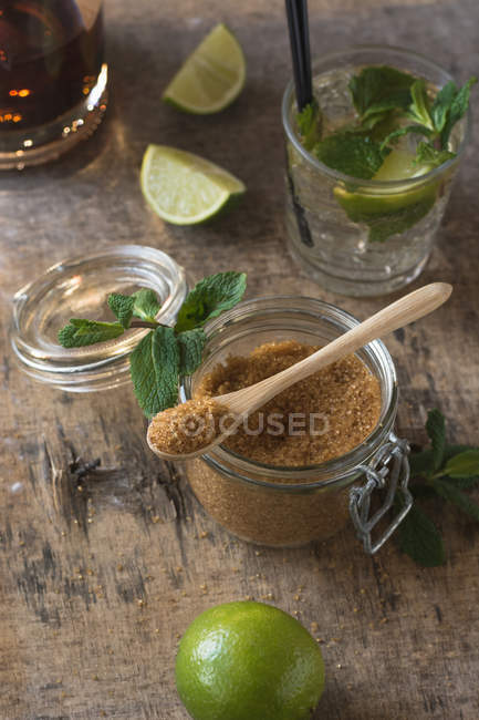 Limas frescas y hojas de menta colocadas en servilleta y mesa cerca de ron y azúcar morena para la preparación de mojitos - foto de stock