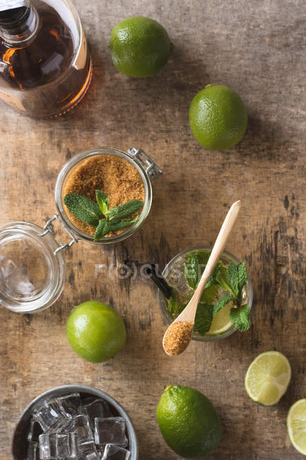 Limas frescas y hojas de menta colocadas en servilleta y mesa cerca de ron y azúcar morena para la preparación de mojitos - foto de stock