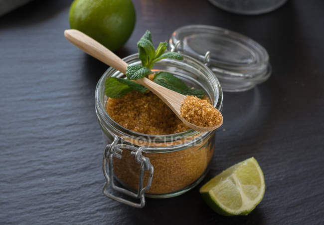 Sucre brun frais dans un bocal près de limes fraîches et de feuilles de menthe poivrée placées sur fond noir — Photo de stock
