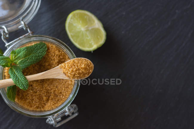 Zucchero di canna in un barattolo vicino a lime fresche e foglie di menta piperita poste su fondo nero — Foto stock