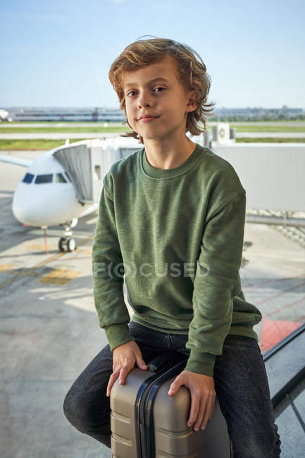 Préadolescent optimiste en vêtements décontractés souriant et assis sur la valise près de la fenêtre dans l'aéroport contemporain — Photo de stock