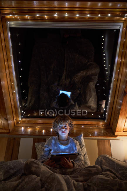 Bambino in pigiama tablet navigazione mentre si trova a letto vicino alla finestra del tetto decorato con luci fata di notte in camera accogliente — Foto stock