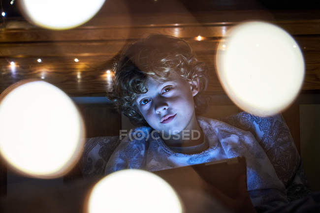 Menino olhando para a câmera e usando tablet enquanto relaxa na cama no quarto acolhedor iluminado com luzes de fadas — Fotografia de Stock