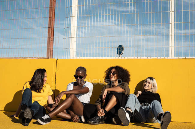 Беззаботные молодые разнообразные женщины в повседневной одежде смеются и дружелюбно беседуют, сидя на спортивной площадке — стоковое фото