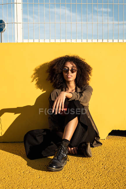 Thoughtful hipster in sunglasses and with Afro hairstyle looking at camera and sitting in sunlight on yellow asphalt on stadium — Stock Photo