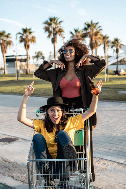 Jeune femme noire en casual et lunettes de soleil debout sur la route à côté du panier tandis que l'autre femme assise à l'intérieur du panier — Photo de stock