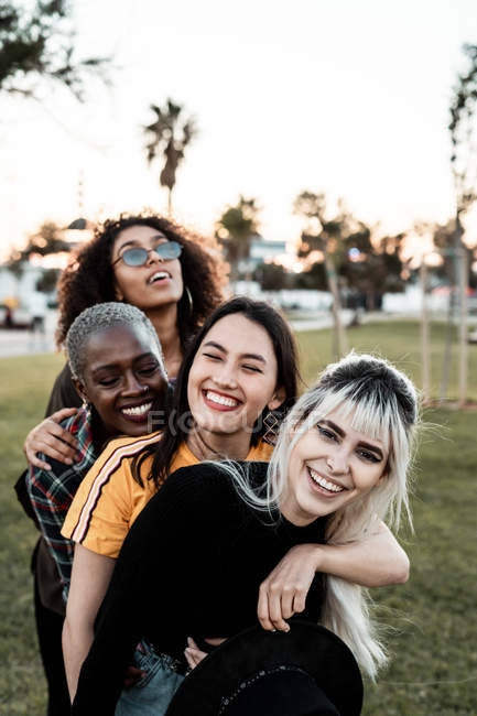 Grupo multiétnico de jóvenes amigas riendo en ropa casual divirtiéndose y abrazándose con cada una en el césped - foto de stock