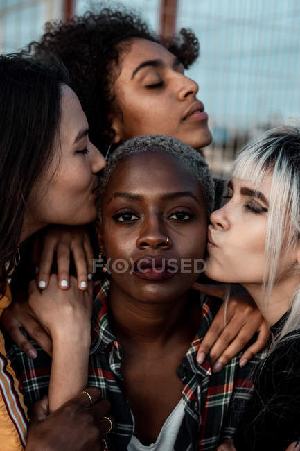 Porträt einer jungen schwarzhaarigen Frau, die mit ernstem Gesicht in die Kamera blickt, während Freundinnen sie mit Umarmungen und Küssen unterstützen — Stockfoto